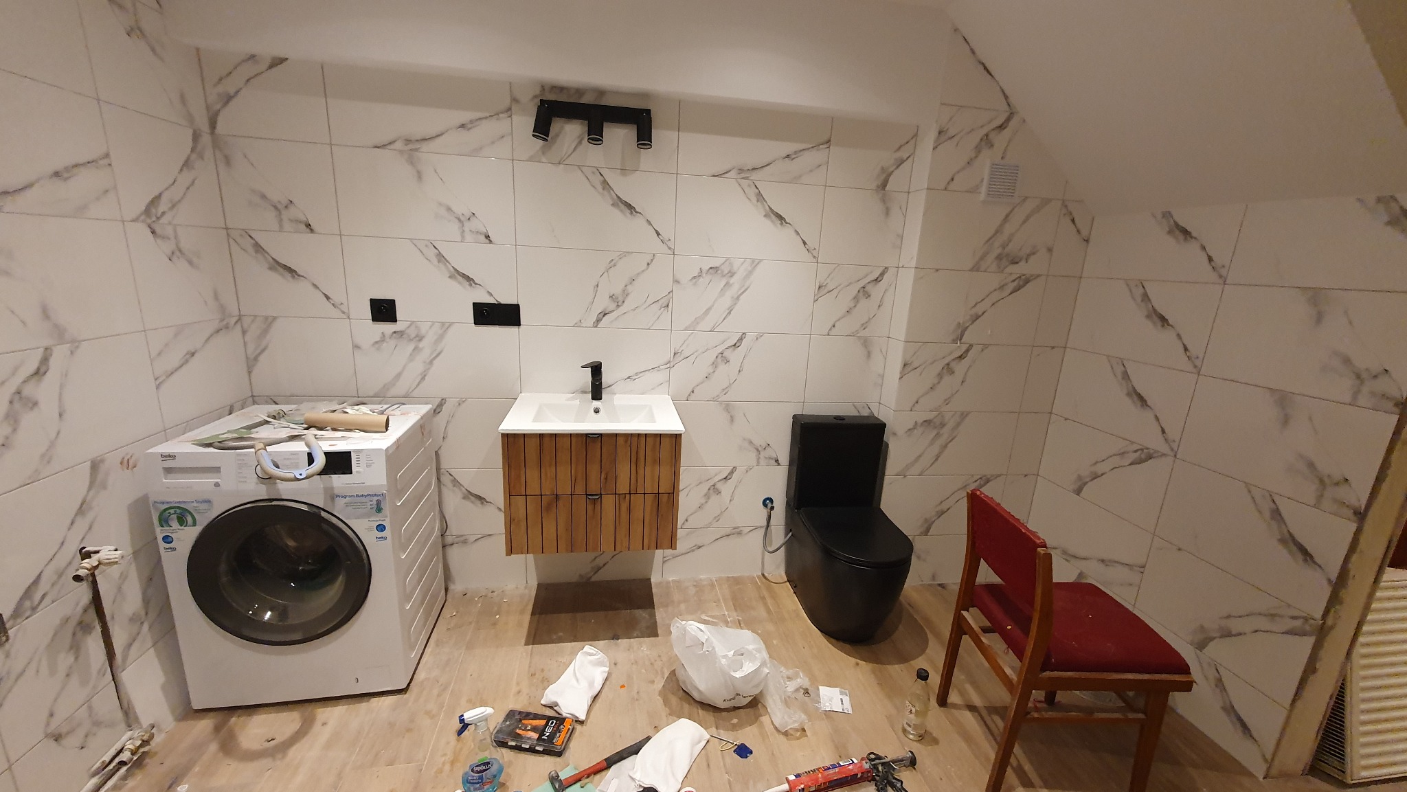 Wykończona łazienka na poddaszu z marmurowymi płytkami, wiszącą szafką z umywalką, czarną toaletą i pralką. Widoczne narzędzia sugerują prace wykończeniowe.