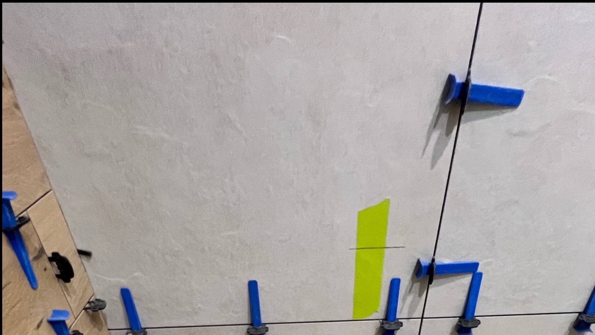 Układanie jasnych płytek z użyciem niebieskich klipsów poziomujących i zielonej taśmy. Widoczne fugi i fragmenty płytek. Detal z prac wykończeniowych.