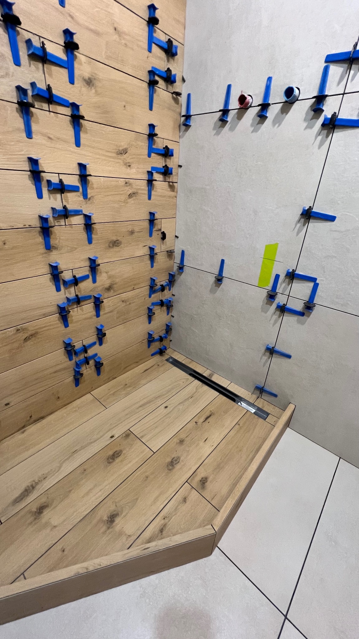 Układanie płytek imitujących drewno w kabinie prysznicowej. Widoczne niebieskie klipsy poziomujące i odpływ liniowy. Jasne płytki na ścianie w tle.