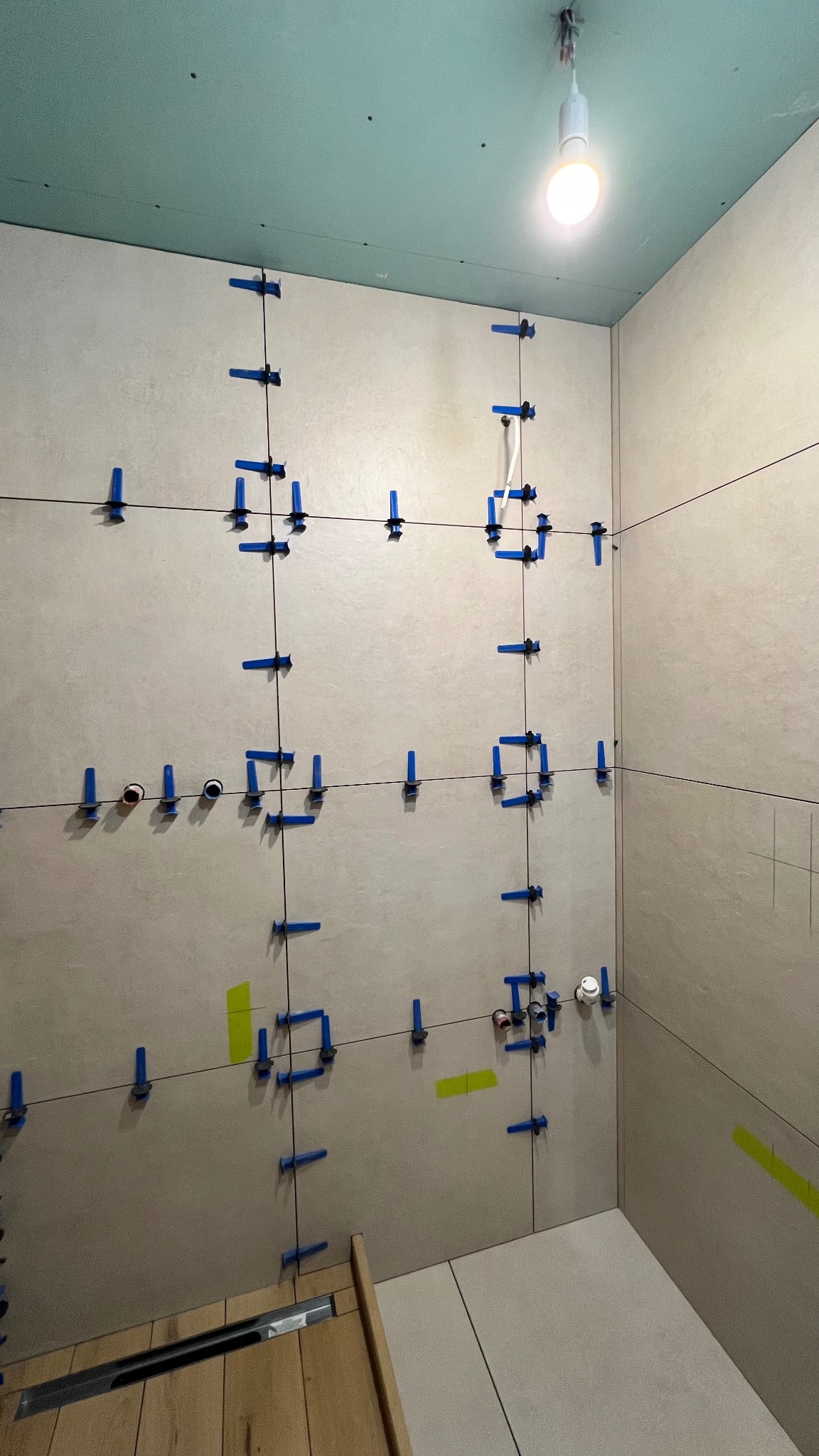 Ściana łazienki w trakcie układania płytek, z użyciem niebieskich krzyżyków dystansowych i poziomicy laserowej. Widoczne punkty instalacji wodnej i fragment odpływu liniowego.