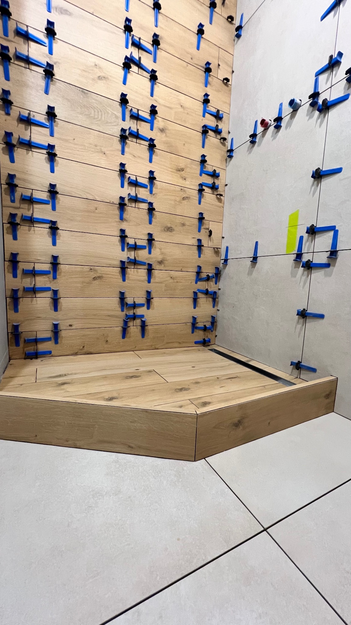 Kąpiel w trakcie wykańczania: drewnopodobne płytki na ścianie, system poziomowania płytek z niebieskimi klipsami, szara ściana w tle. Widoczny odpływ liniowy.