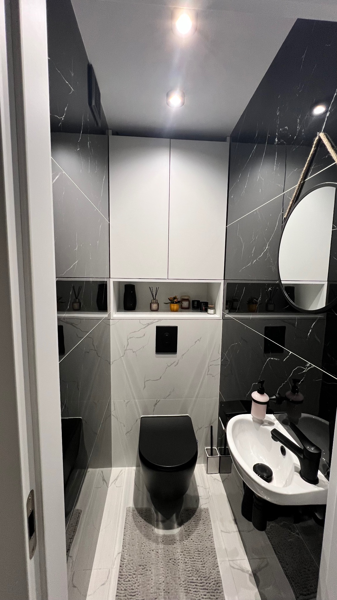 Nowoczesna łazienka w czerni i bieli z marmurowymi płytkami, podwieszaną toaletą i okrągłym lustrem na sznurze. Minimalistyczny design z akcentami dekoracyjnymi na półkach.