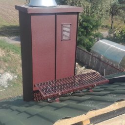 Bartosz Tomczak - Z bliska: brązowy komin z metalową drabinką na zielonym dachu z blachy falistej. W tle widoczny ogródek szklarniowy i pagórek z drewnem.