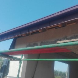 Bartosz Tomczak - Niski kąt ujęcia na brązową rynnę i fragment dachu z rusztowaniem. Widoczne elementy konstrukcyjne i fragment elewacji budynku w trakcie prac.