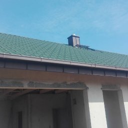 Bartosz Tomczak - Zielony dach z brązowymi rynnami i kominem na tle błękitnego nieba. Widoczne surowe ściany budynku w stanie deweloperskim.