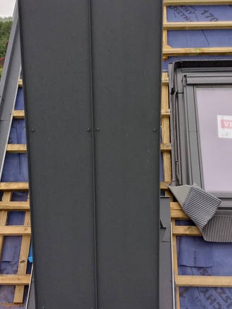 Montaż paneli dachowych z oknem dachowym Velux. Widoczne drewniane łaty i membrana dachowa. Detal dachu w trakcie budowy lub remontu.