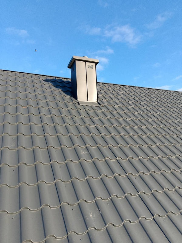 Szary dach z blachodachówki z kominem w słoneczny dzień. Widoczny fragment nieba z chmurami i przelatujący ptak. Komin w kolorze dachu.