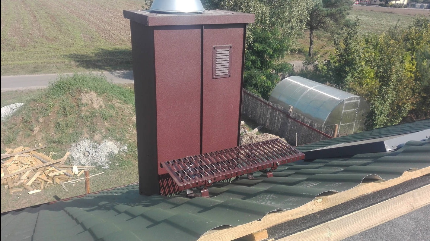 Z bliska: brązowy komin z metalową drabinką na zielonym dachu z blachy falistej. W tle widoczny ogródek szklarniowy i pagórek z drewnem.