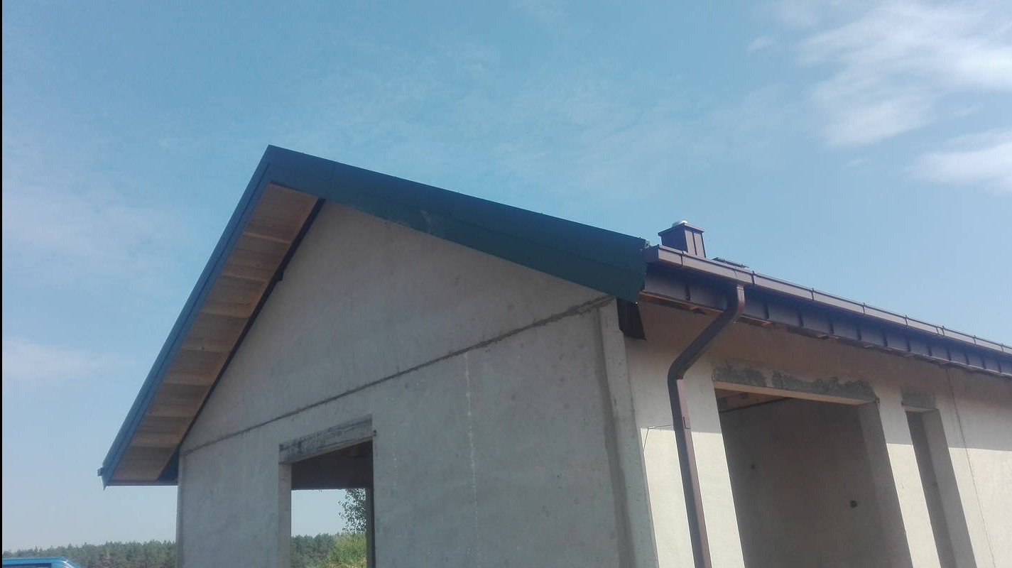 Surowy stan domu z widoczną konstrukcją dachu i rynnami w kolorze brązowym na tle błękitnego nieba. Widoczne elementy drewniane i betonowe.