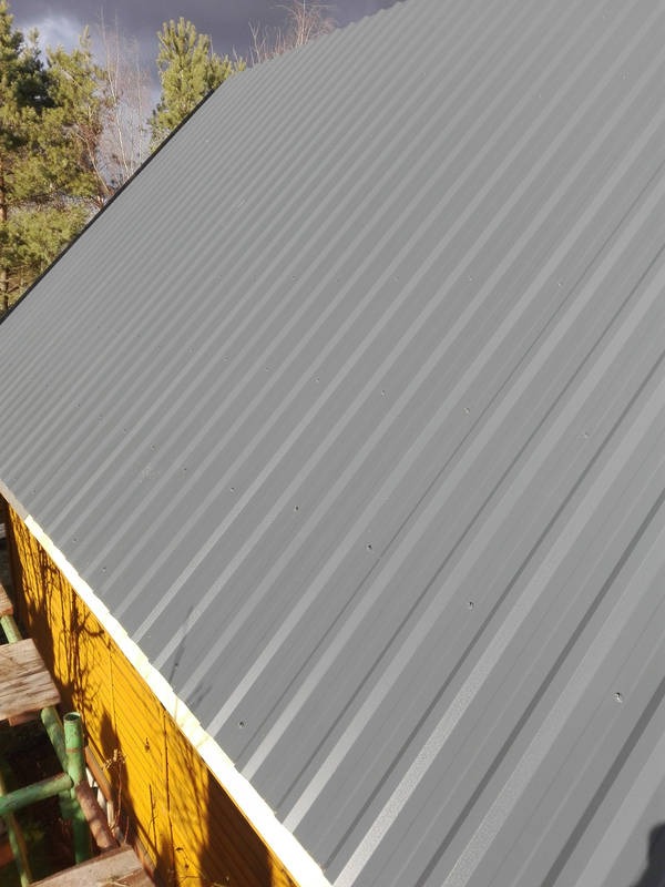 Szary dach z blachy trapezowej, widok z dołu pod kątem. Żółta ściana budynku i fragment rusztowania w dolnej części kadru. Pochmurne niebo w tle.