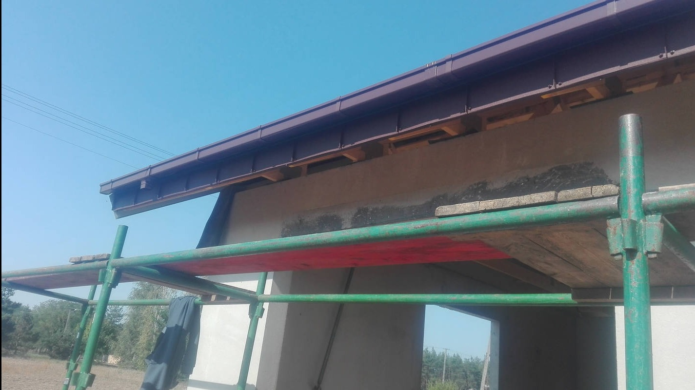 Niski kąt ujęcia na brązową rynnę i fragment dachu z rusztowaniem. Widoczne elementy konstrukcyjne i fragment elewacji budynku w trakcie prac.