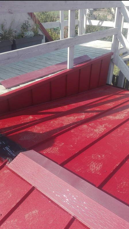 Czerwony dach z blachy trapezowej w trakcie krycia, widoczne ślady stóp, fragment membrany dachowej i biała balustrada w tle. Ujęcie z góry.