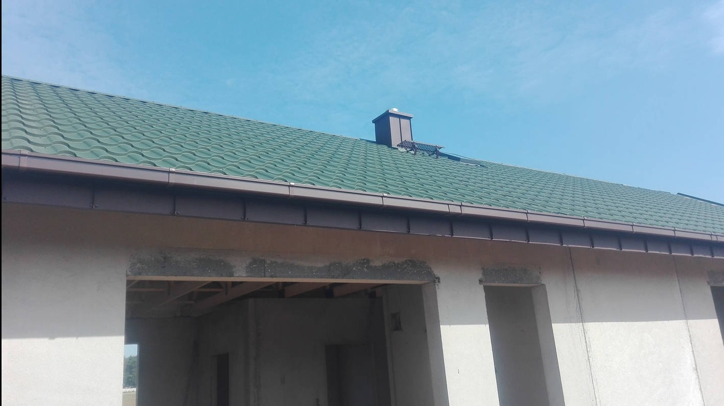 Zielony dach z brązowymi rynnami i kominem na tle błękitnego nieba. Widoczne surowe ściany budynku w stanie deweloperskim.