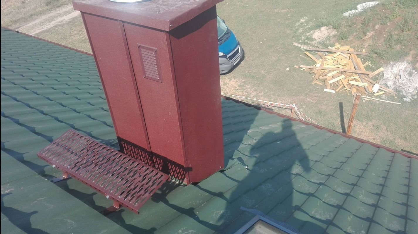 Zielony dach z blachy trapezowej, brązowy komin wentylacyjny z kratką, metalowa ława kominiarska. W tle sterta drewna i fragment samochodu.