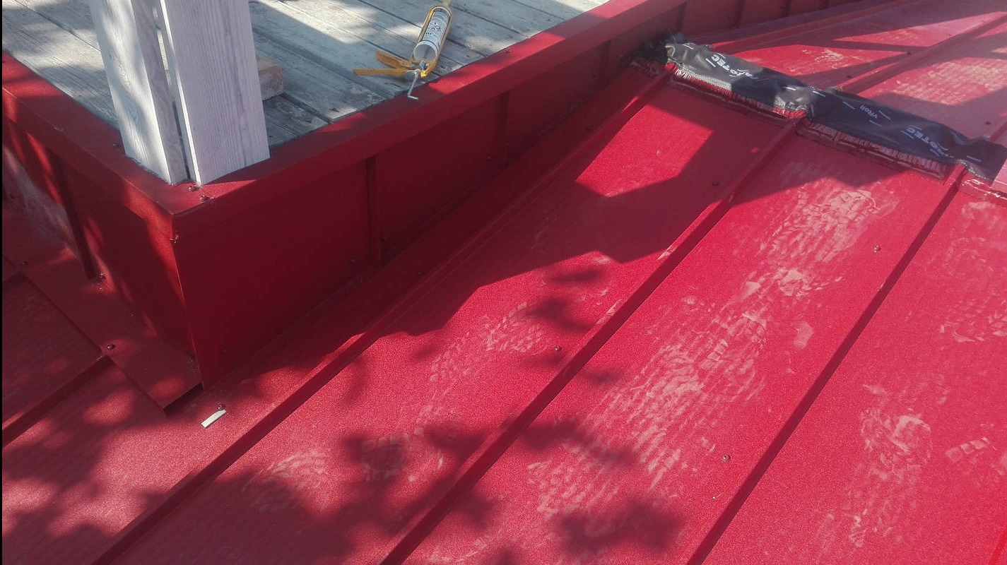 Czerwony dach z blachy trapezowej, widoczne obróbki blacharskie przy kominie i narzędzia dekarskie. Ślady butów na powierzchni dachu.