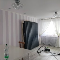 Ewa Wichrowska - Pokój po malowaniu ścian w pionowe pasy, z materacem opartym o ścianę, białym stołem, krzesłem i odkurzaczem. Dwa kryształowe żyrandole.