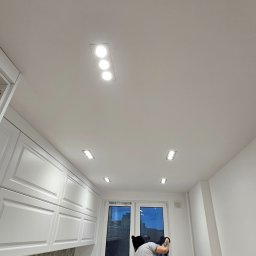 Ewa Wichrowska - Montaż oświetlenia sufitowego w kuchni: mężczyzna instaluje lampy w białym suficie, widoczne białe szafki kuchenne i okno. Ujęcie z dołu, podkreślające oświetlenie.