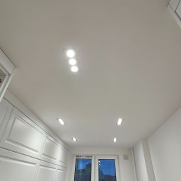 Ewa Wichrowska - Biała kuchnia po remoncie z oświetleniem sufitowym. Mężczyzna sprząta blat po montażu. Widok na okno z odbiciem budynku. Minimalistyczny design.