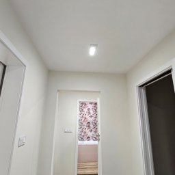 Ewa Wichrowska - Korytarz po malowaniu na biało z widokiem na pokój z tapetą w kwiaty. Widoczne listwy przypodłogowe i oświetlenie sufitowe. Podłoga zabezpieczona kartonem.