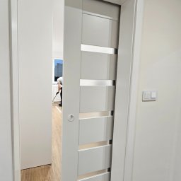 DO4U remonty - Białe, przesuwne drzwi z poziomymi, srebrnymi wstawkami, częściowo otwarte, ukazujące wnętrze z osobą sprzątającą. Widoczny fragment podłogi i włącznik światła.