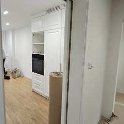 DO4U remonty - Biała zabudowa kuchenna z piekarnikiem w otwartej przestrzeni, widoczna zza białych drzwi przesuwnych. Na podłodze panele imitujące drewno. Rolka materiału na podłodze.