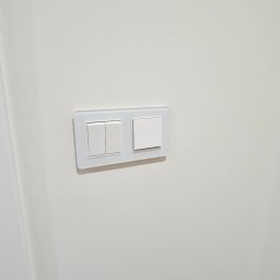 DO4U remonty - Dwa białe, nowoczesne włączniki światła z przezroczystą ramką, zamontowane na gładkiej, białej ścianie. Minimalistyczny design, oświetlenie z góry.