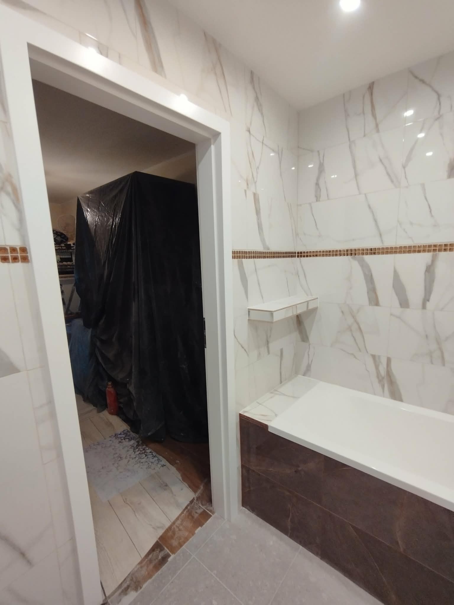 Remont łazienki w trakcie: biało-brązowe płytki imitujące marmur na ścianach, obudowana wanna, półka. W tle zabezpieczone folią meble i podłoga w remoncie.