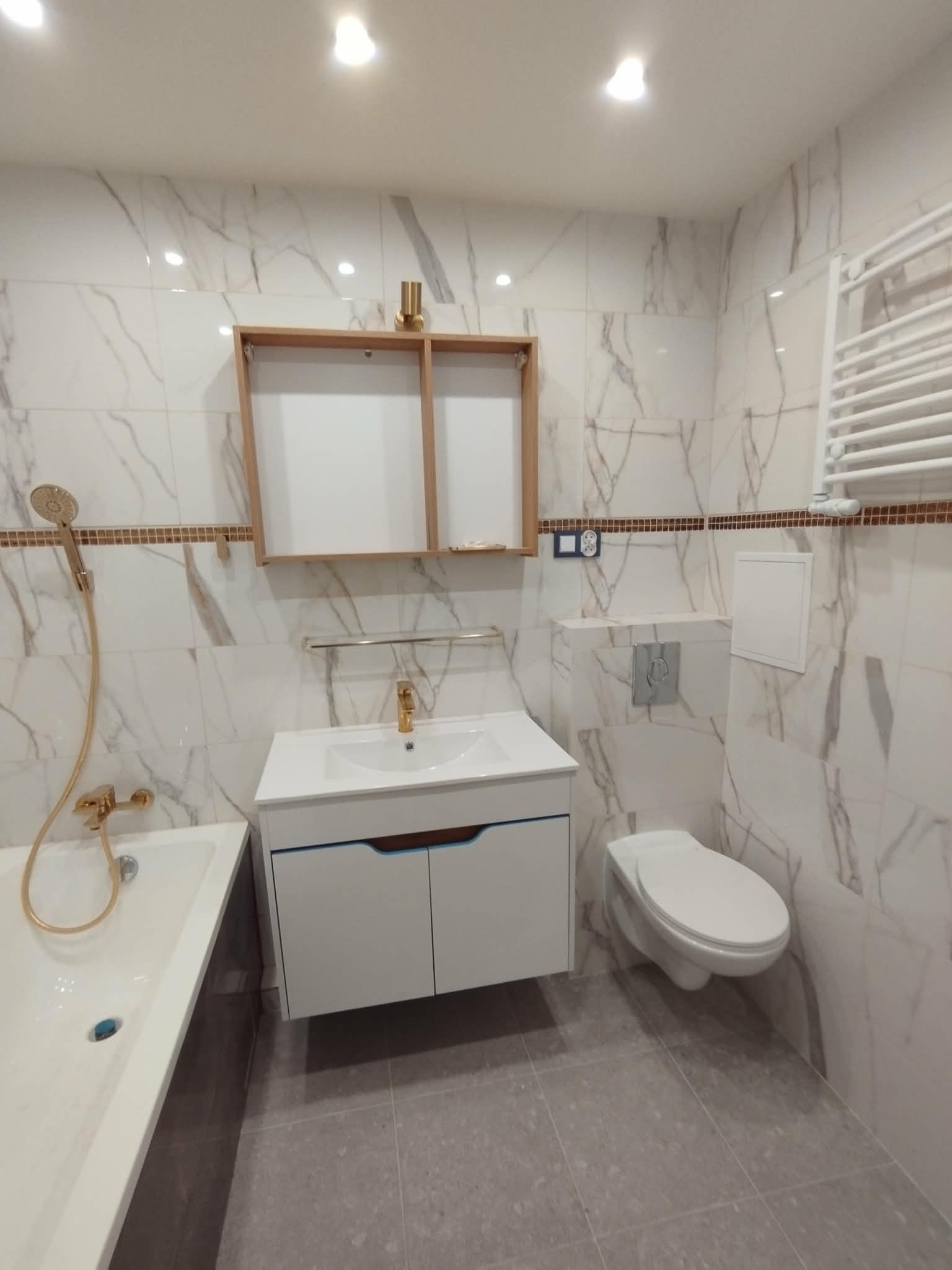 Wykończona łazienka z marmurowymi płytkami, wiszącą szafką z umywalką, toaletą podwieszaną i grzejnikiem drabinkowym. Złota armatura dodaje elegancji.