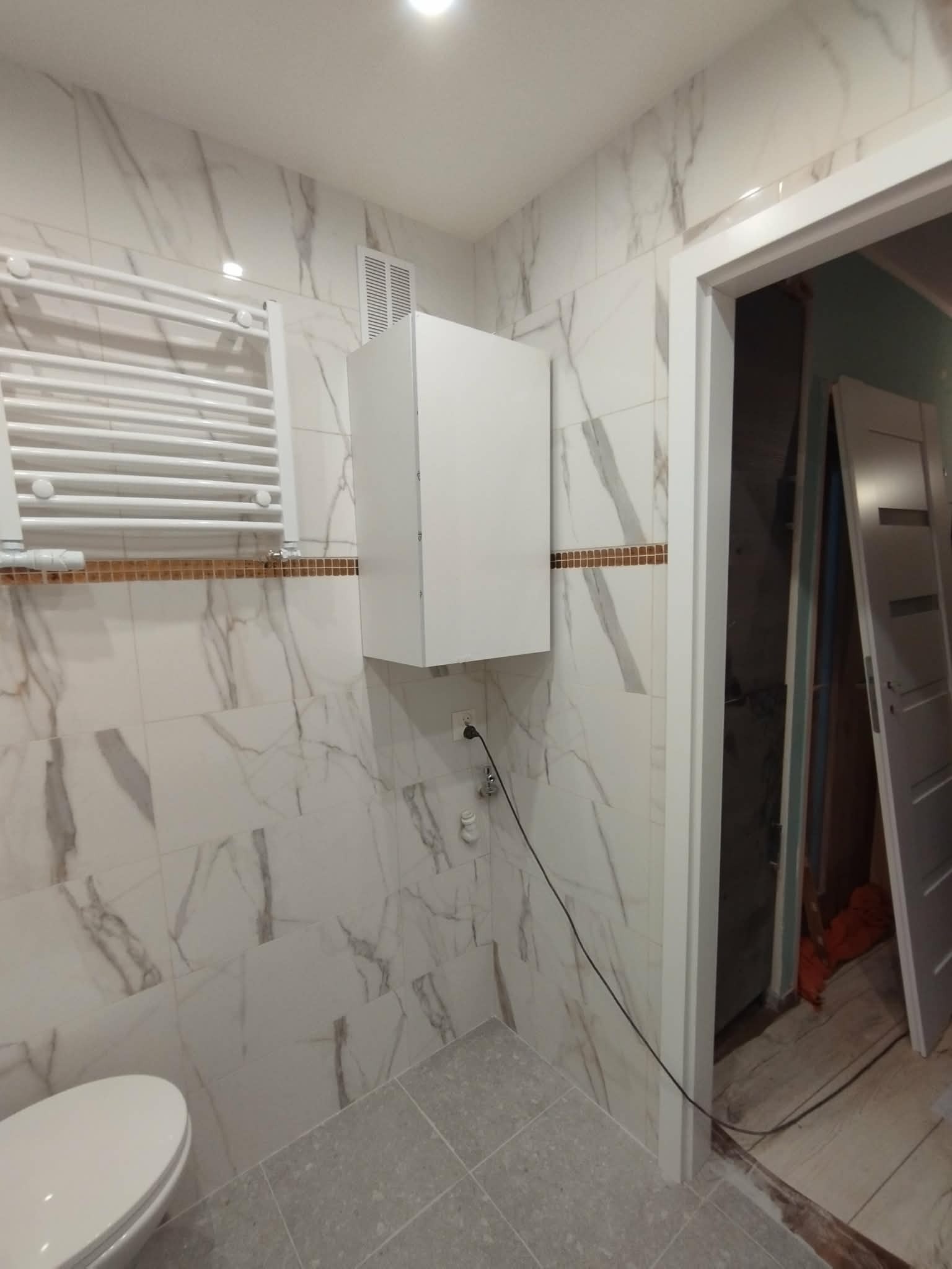 Wykończona łazienka z marmurowymi płytkami, grzejnikiem drabinkowym, szafką wiszącą i częściowo widoczną toaletą. Widoczne niedokończone obramowanie drzwi.