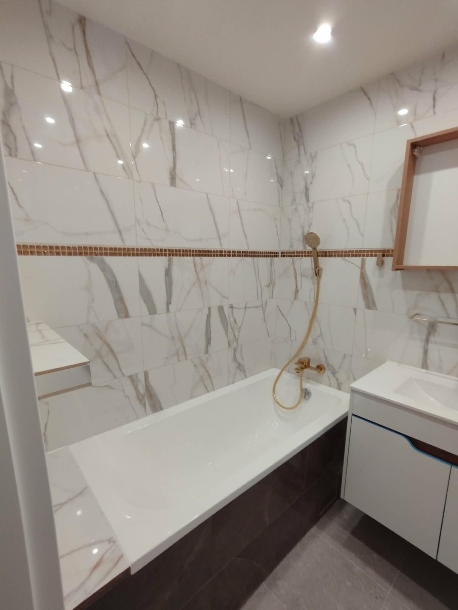 Nowoczesna łazienka po remoncie: białe marmurowe płytki z brązowymi żyłkami, wanna z brązową obudową, biała szafka z umywalką i złota armatura.