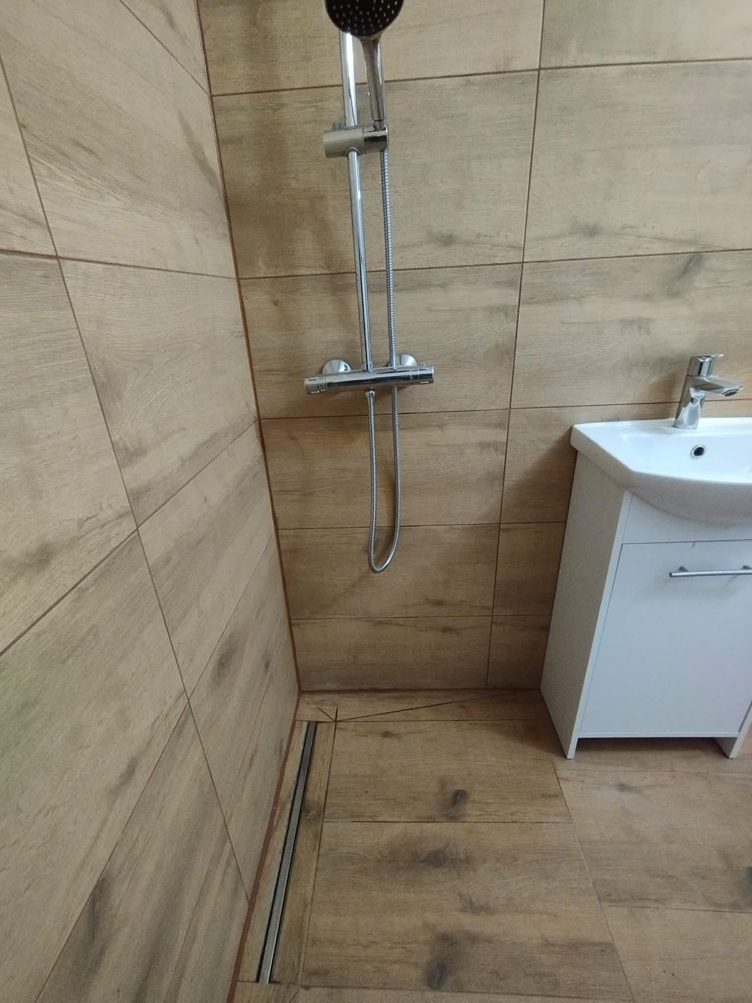 Nowoczesna kabina prysznicowa z odpływem liniowym i umywalką. Płytki imitujące drewno na ścianach i podłodze. Chromowana armatura. Minimalistyczny design.