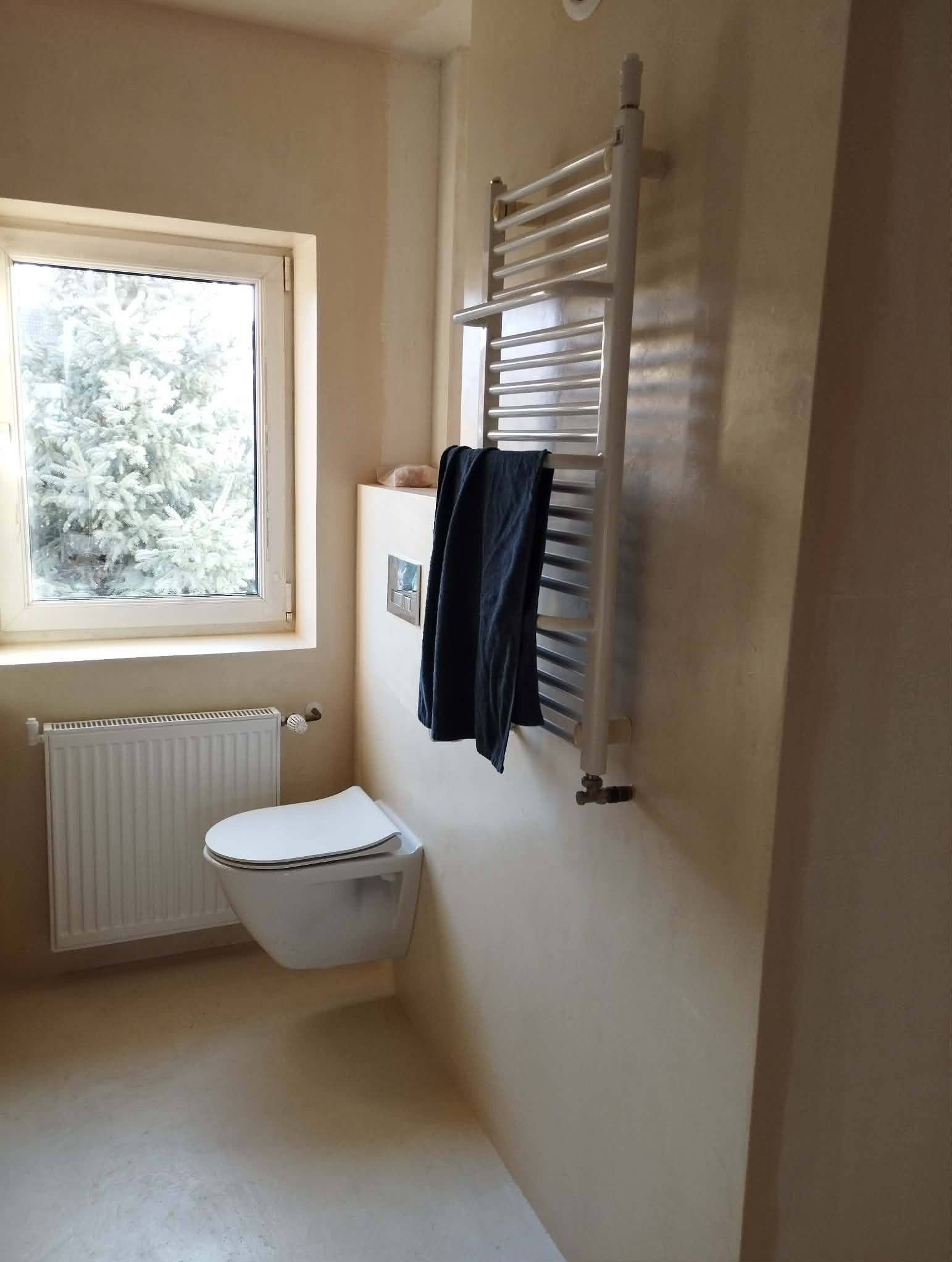 Jasna łazienka po remoncie: podwieszana toaleta, grzejnik drabinkowy z ręcznikiem, okno z widokiem na drzewo. Ściany w odcieniach beżu, minimalistyczny styl.