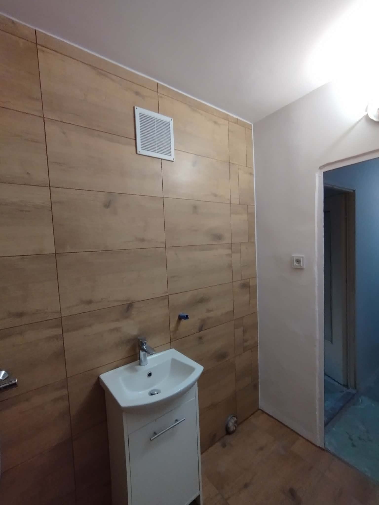 Wykończona łazienka z drewnopodobnymi płytkami, białą szafką z umywalką i wentylatorem. Widoczny fragment drzwi do kabiny prysznicowej.