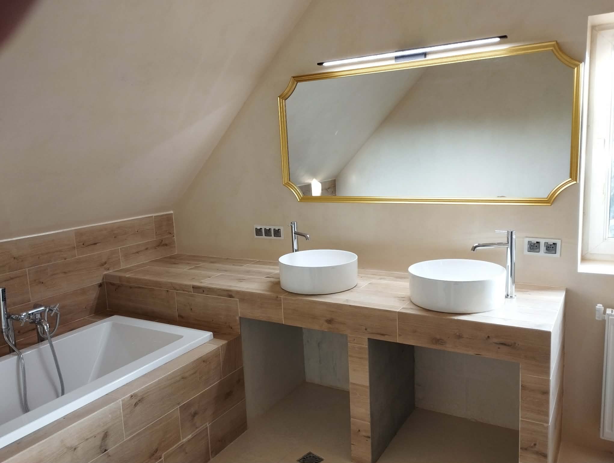 Wykończona łazienka na poddaszu z dwiema okrągłymi umywalkami, lustrem w złotej ramie i wanną obudowaną płytkami imitującymi drewno. Ukośne ściany.