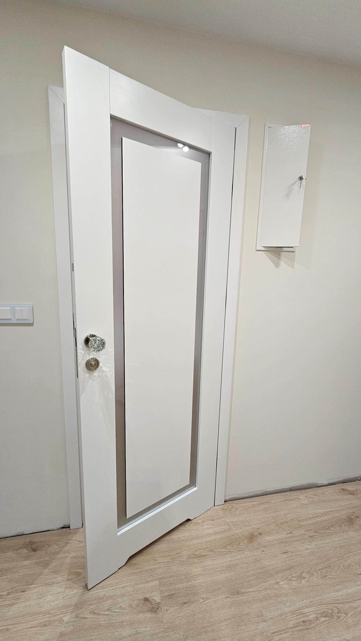 Białe, częściowo otwarte drzwi z przeszkleniem i kryształową gałką na tle jasnej ściany i podłogi imitującej drewno. Widoczna skrzynka elektryczna.