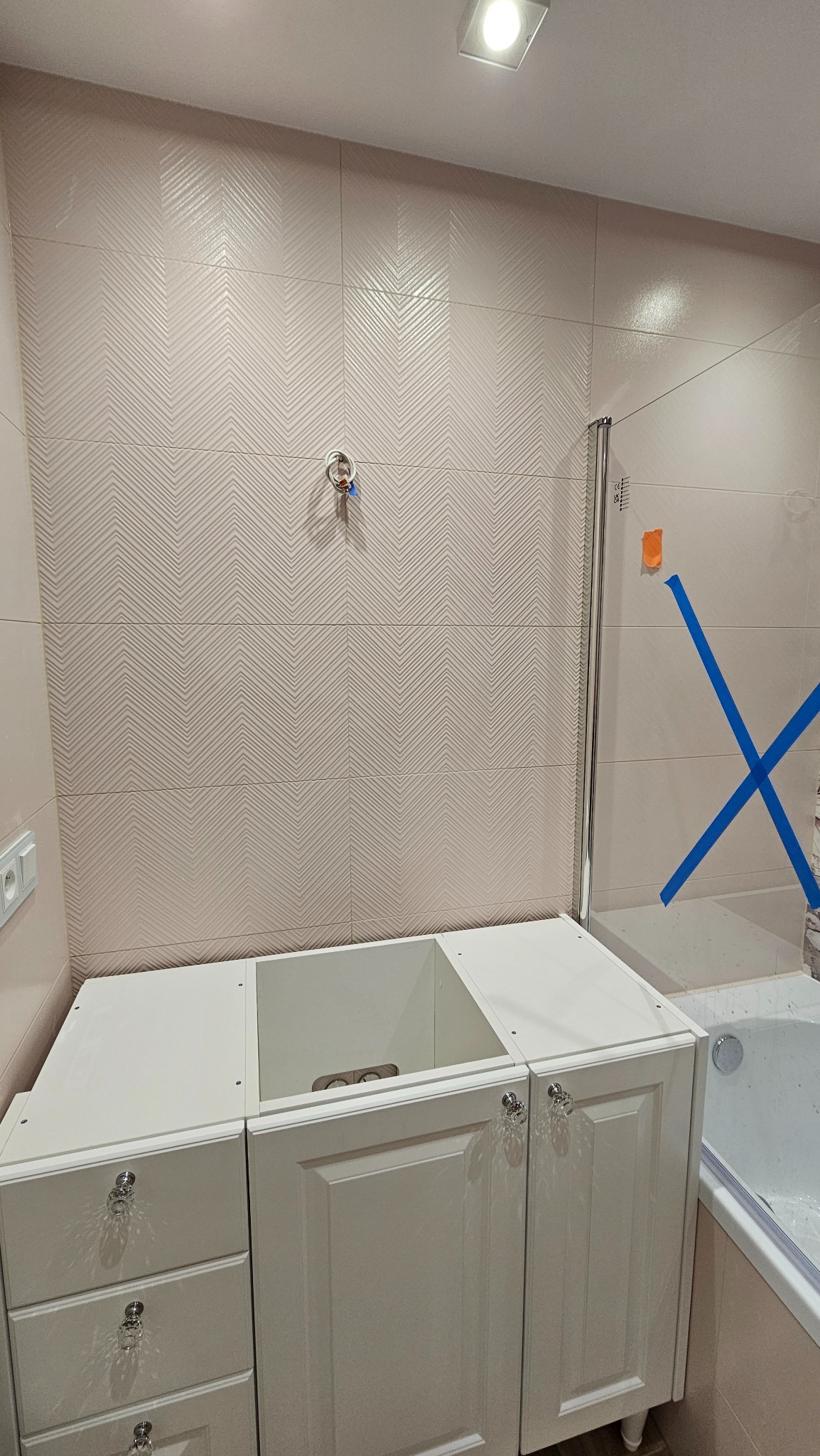 Biała szafka pod umywalkę z kryształowymi uchwytami na tle beżowych płytek w jodełkę i częściowo widocznej kabiny prysznicowej z niebieską taśmą.