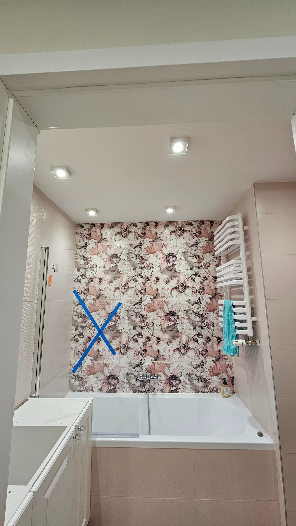 Wykończona łazienka z wanną, grzejnikiem drabinkowym i kwiatową tapetą. Kabina prysznicowa z niebieskim krzyżem. Widok zza framugi drzwi.