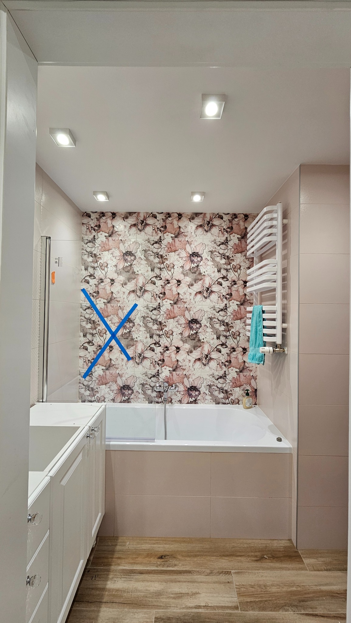 Jasna łazienka z wanną, grzejnikiem drabinkowym i umywalką w trakcie montażu. Na ścianie dekoracyjne płytki z motywem kwiatowym, zabezpieczone taśmą.