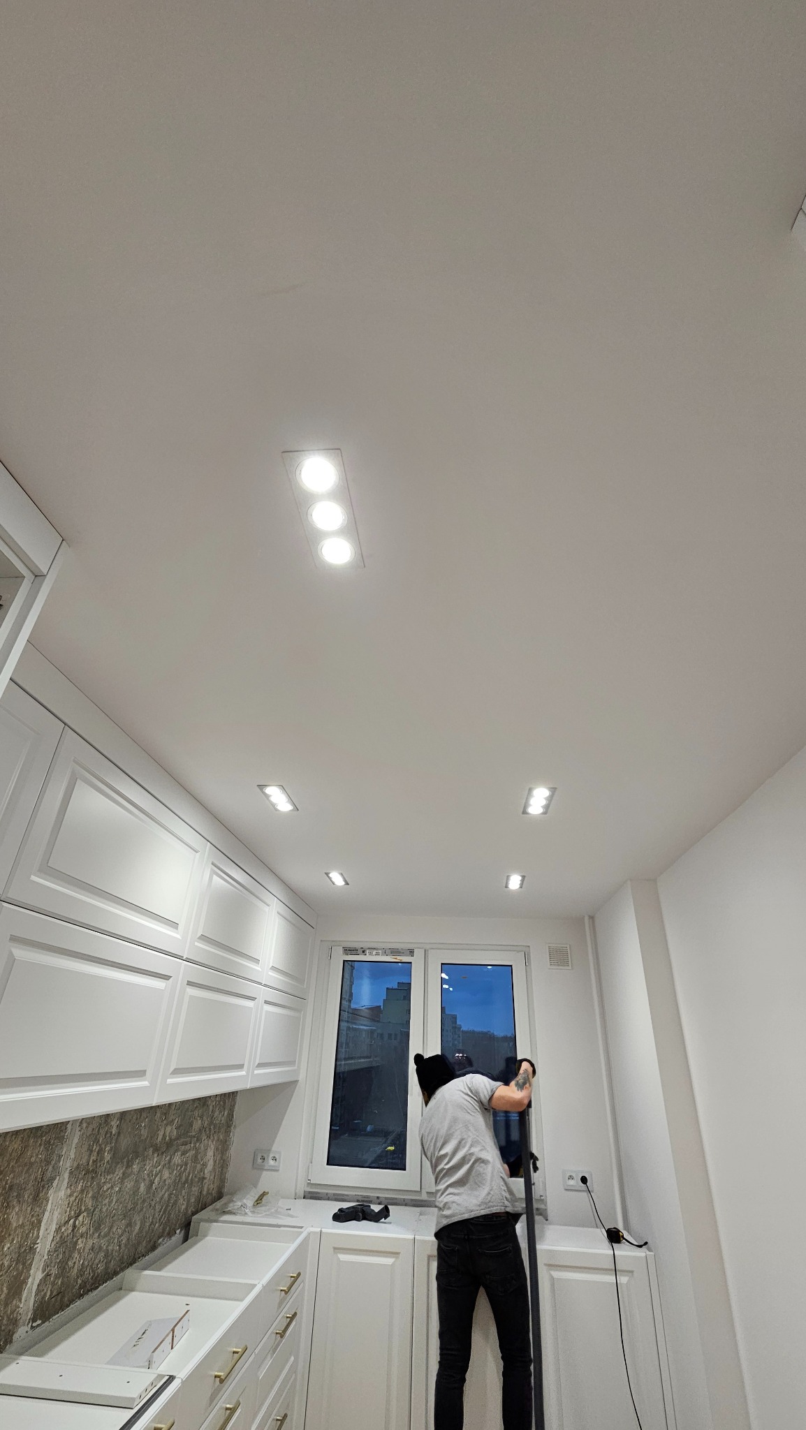 Montaż oświetlenia sufitowego w kuchni: mężczyzna instaluje lampy w białym suficie, widoczne białe szafki kuchenne i okno. Ujęcie z dołu, podkreślające oświetlenie.