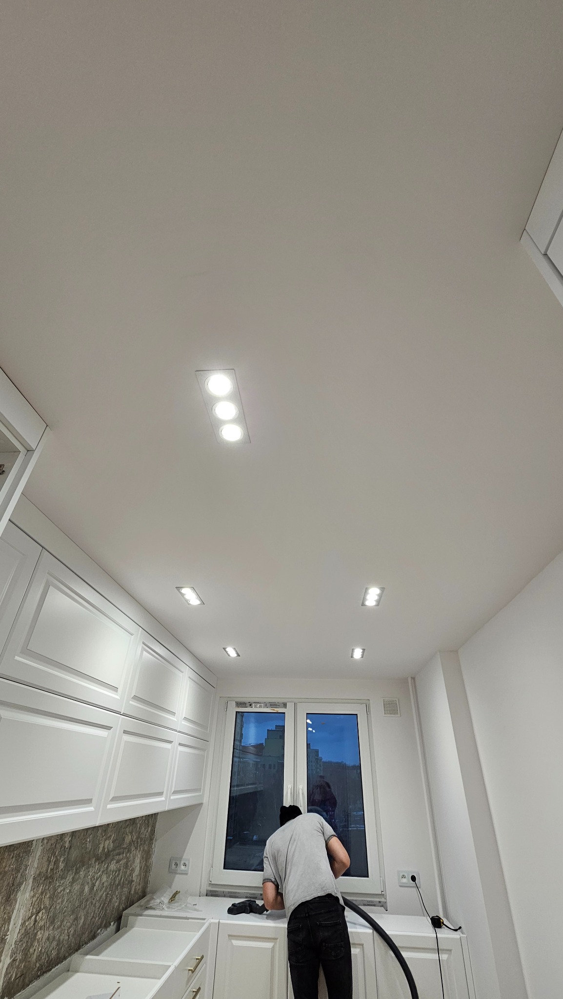 Biała kuchnia po remoncie z oświetleniem sufitowym. Mężczyzna sprząta blat po montażu. Widok na okno z odbiciem budynku. Minimalistyczny design.