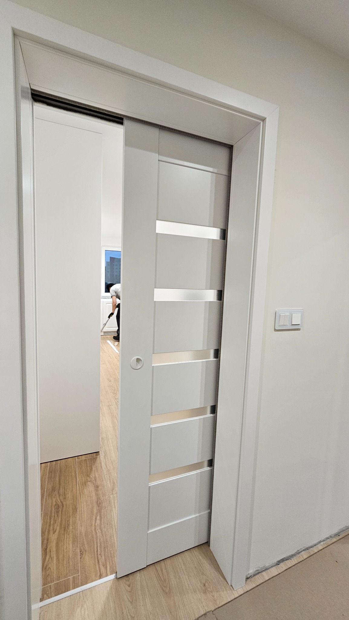 Białe, przesuwne drzwi z poziomymi, srebrnymi wstawkami, częściowo otwarte, ukazujące wnętrze z osobą sprzątającą. Widoczny fragment podłogi i włącznik światła.