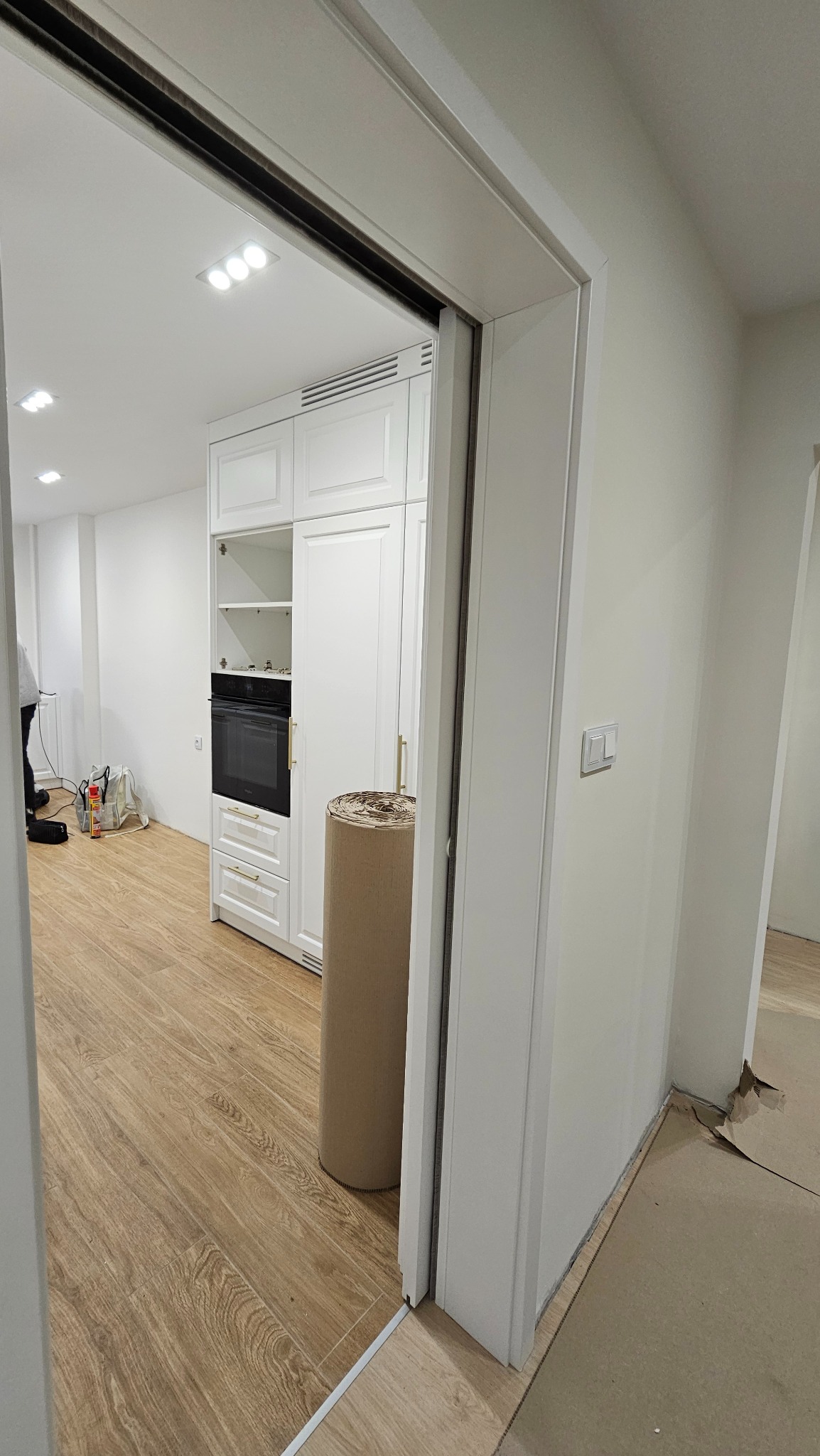 Biała zabudowa kuchenna z piekarnikiem w otwartej przestrzeni, widoczna zza białych drzwi przesuwnych. Na podłodze panele imitujące drewno. Rolka materiału na podłodze.