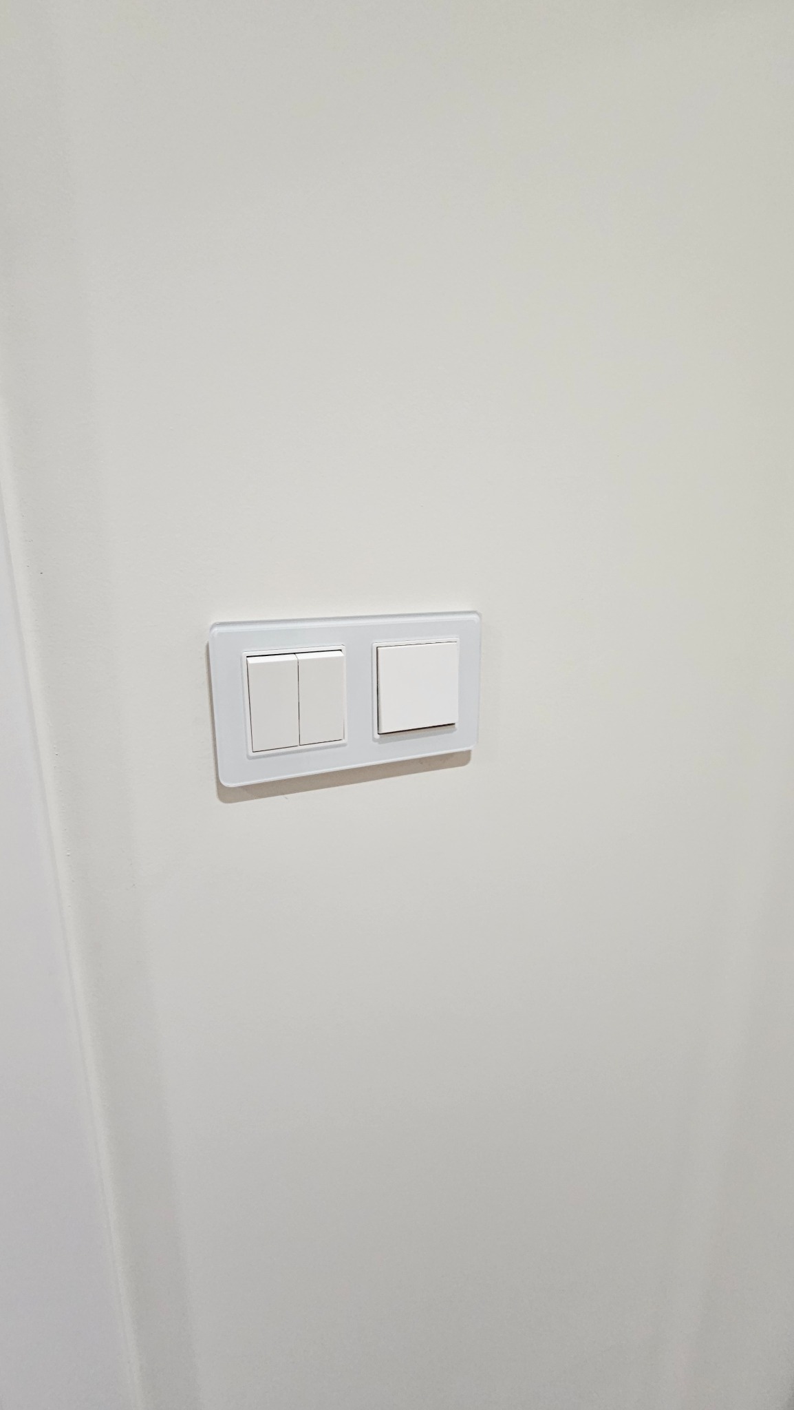 Dwa białe, nowoczesne włączniki światła z przezroczystą ramką, zamontowane na gładkiej, białej ścianie. Minimalistyczny design, oświetlenie z góry.