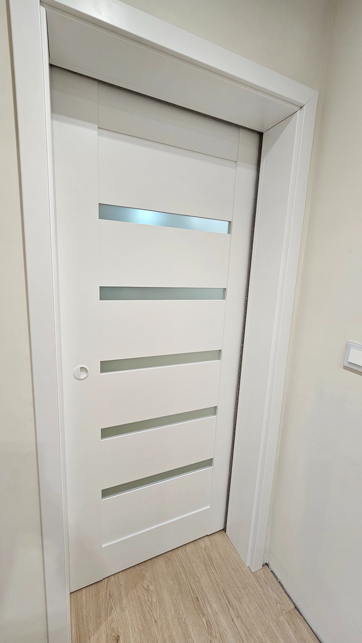 Białe drzwi wewnętrzne z poziomymi przeszkleniami, zamontowane w ościeżnicy. Widoczna podłoga z paneli i fragment ściany z włącznikiem.