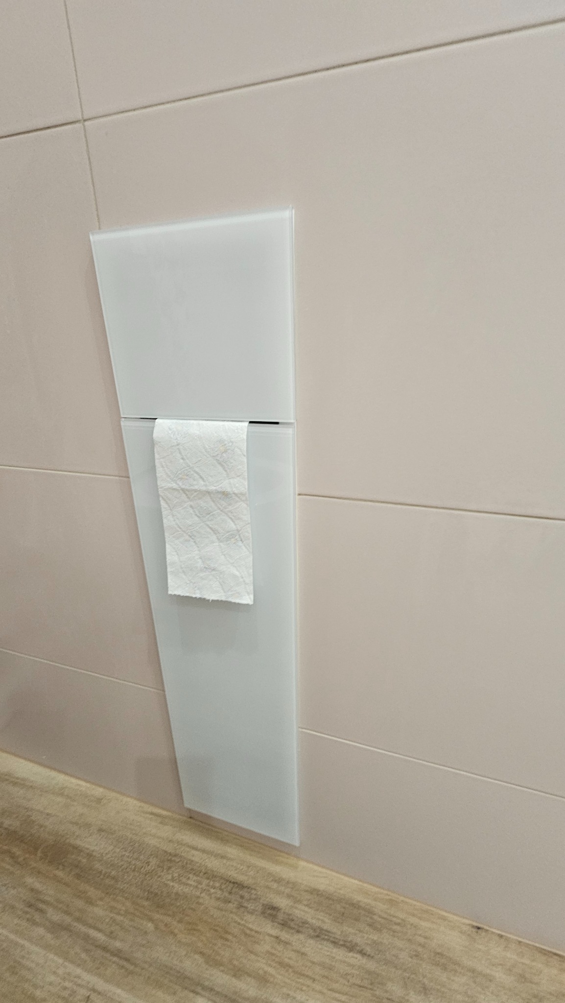 Biały, szklany podajnik na papier toaletowy wkomponowany w beżowe płytki ścienne, z rolką papieru w środku. Widoczny fragment drewnianej podłogi.