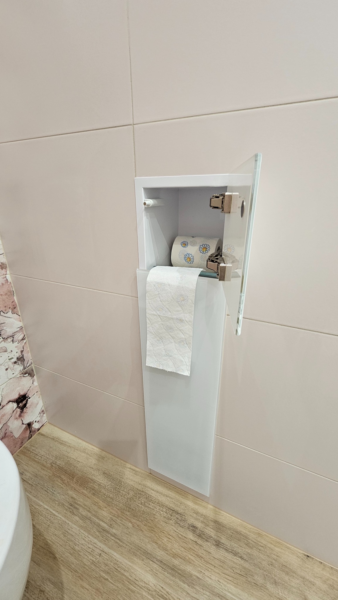 Wnęka w łazience z ukrytym pojemnikiem na papier toaletowy, wykończona beżowymi płytkami. Widoczny fragment podłogi imitującej drewno i fragment muszli klozetowej.