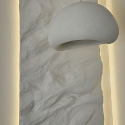Studio Projektowania Wnętrz MONIKA STAWOWCZYK - Biała lampa wisząca o organicznym kształcie na tle ściany z dekoracyjną, teksturowaną fakturą i subtelnym oświetleniem LED. Minimalistyczny design.