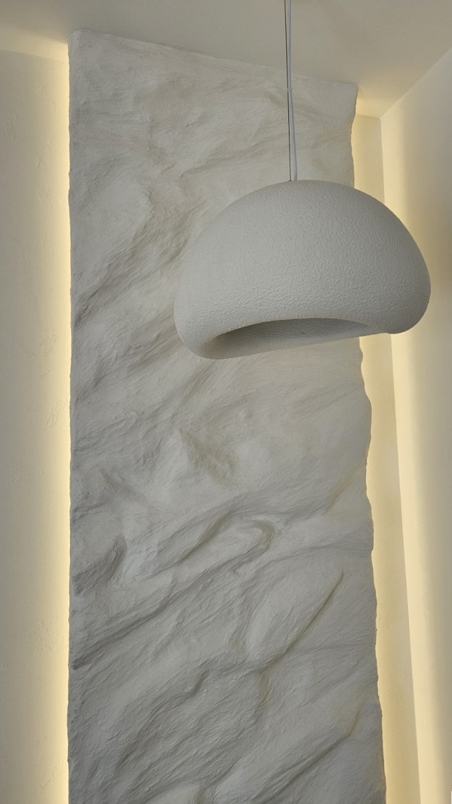 Biała lampa wisząca o organicznym kształcie na tle ściany z dekoracyjną, teksturowaną fakturą i subtelnym oświetleniem LED. Minimalistyczny design.