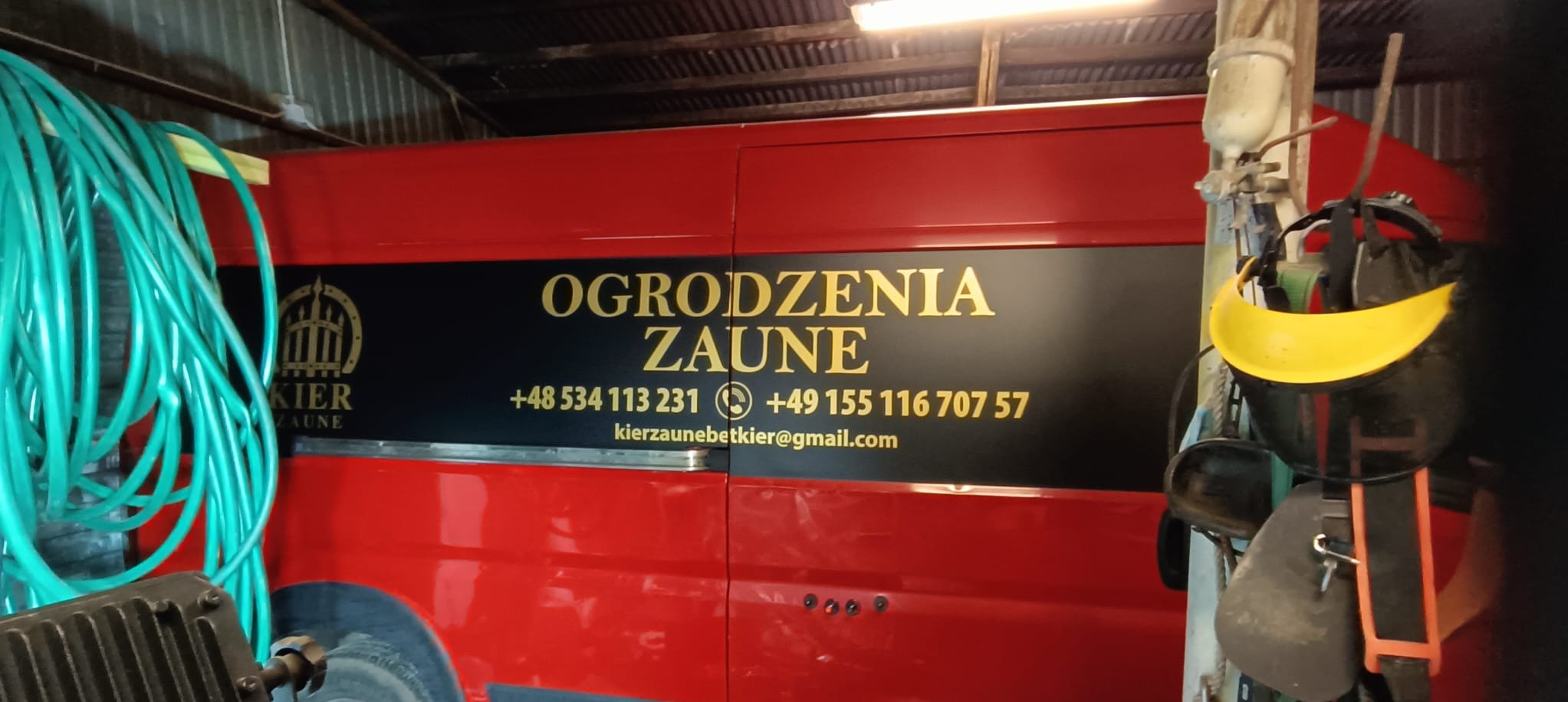Czerwony samochód dostawczy z logo firmy 'Ogrodzenia Zaune' i numerami telefonów, zaparkowany w warsztacie z narzędziami i wężem ogrodowym w tle.