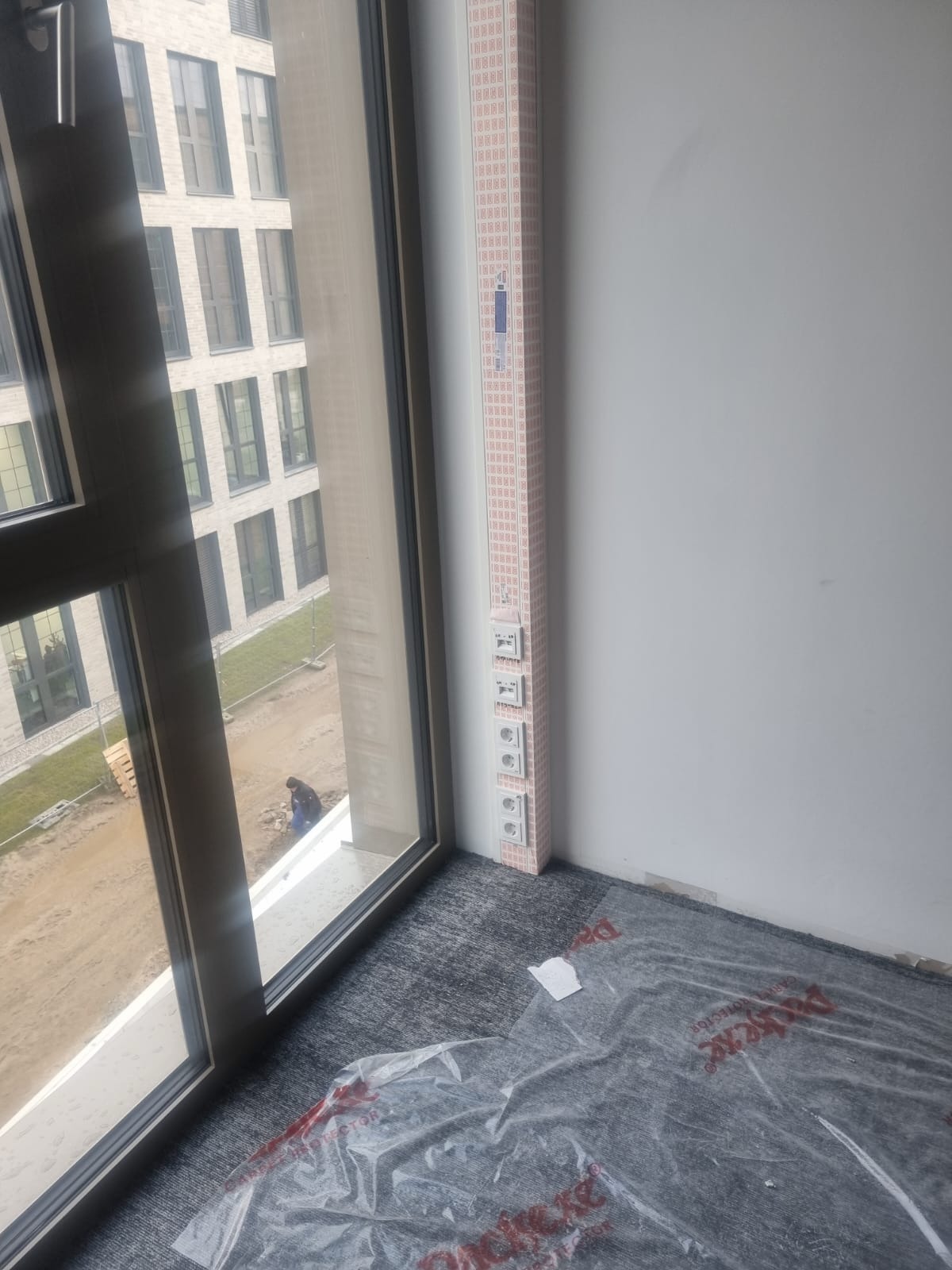 Nowe gniazdka elektryczne zamontowane w pionowej listwie przy oknie w jasnym pokoju. Podłoga zabezpieczona folią ochronną. Widok na budynek z zewnątrz.